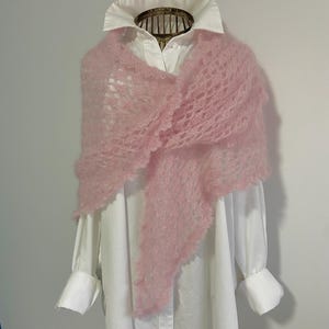 Peut inclure: Un châle triangulaire rose clair en tricot ajouré à l'aspect duveteux et texturé, drapé sur une chemise blanche à boutons. Le châle présente un motif délicat en dentelle sur toute sa surface avec un détail festonné le long du bord.