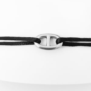 Könnte beinhalten: Ein schwarzes Kordelarmband mit einem silbernen Metallverschluss in Form eines Schiffsankers.