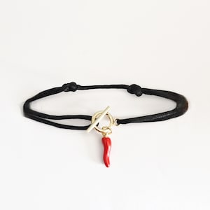 Peut inclure: Bracelet en cordon noir avec un fermoir à bascule doré et un charm en forme de piment rouge. Le bracelet est présenté sur fond blanc. Le charm est de couleur rouge vif.