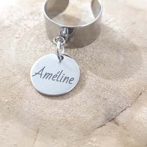 Peut inclure: Une bague réglable argentée avec un charm circulaire. Le charm est gravé du nom "Améline" en écriture cursive. La bague est présentée sur une surface en bois.