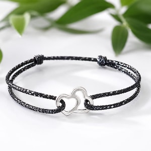 Op de afbeelding: Zwart koordarmband met een zilveren bedel met twee in elkaar grijpende harten. De armband heeft een verstelbare knoopsluiting. De achtergrond is wit met groen blad.