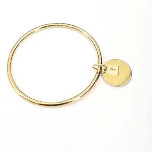 Könnte beinhalten: Ein goldfarbenes Armreif-Armband mit kreisförmigem Design. Ein kleiner, runder Anhänger mit Schlüsselloch-Design hängt am Armband. Das Armband besteht aus glänzendem, metallischem Material und ist vor einem weißen Hintergrund platziert.