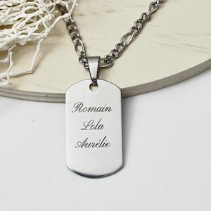 Puede incluir: Un collar con colgante de placa de identificación de color plateado con los nombres "Romain", "Lola" y "Aurélie" grabados. El colgante cuelga de una cadena plateada. El fondo es una superficie de madera clara.