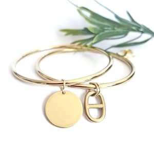 Peut inclure: Deux bracelets dorés avec des breloques. L'un des bracelets a un charme rond, et l'autre a un charme avec un design incurvé. Les bracelets sont présentés sur une surface blanche avec une plante verte en arrière-plan.