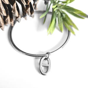 Op de afbeelding: Een zilverkleurige armband met een kleine, ovaalvormige bedel. De bedel heeft een gestileerd ontwerp. De armband wordt gepresenteerd met een dennenappel en een groene tak op een witte achtergrond. Sieraden.