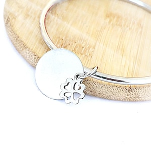 Op de afbeelding: Een zilverkleurige armband met een ronde bedel en een klavervormige bedel. De armband wordt gepresenteerd op een lichtbruin houten oppervlak. De bedels zijn met kleine lusjes aan de armband bevestigd.