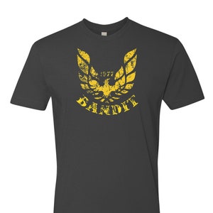 May include: A dark gray t-shirt with a yellow distressed graphic of a bird with wings spread and the text "1977 BANDIT".