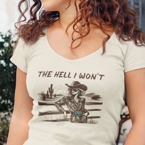 May include: Cream-colored V-neck t-shirt with the text "THE HELL I WON'T" above a graphic of a cowgirl leaning on a fence. The cowgirl is wearing a hat and sunglasses. The background features a desert landscape.