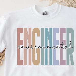 Peut inclure: Un sweat-shirt blanc à manches longues et col rond avec le mot ENGINEER écrit en grandes lettres verticales multicolores. Le mot environmental est superposé au centre en écriture cursive noire fine.