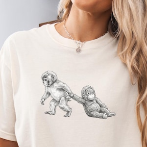 May include: Ivory-colored t-shirt featuring a black and white illustration of two monkeys. One monkey is standing and holding the hand of another monkey that is sitting down. The shirt is a relaxed fit.