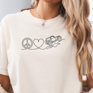 May include: Ivory-coloured t-shirt with a black line drawing of a peace sign, a heart, and a jeep driving over a hill. The shirt has a relaxed fit. The word "Ivory" is printed in the bottom right corner.