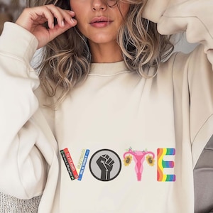 Op de afbeelding: Een wit sweatshirt met het woord "VOTE" gespeld in kleurrijke graphics. De letters zijn gemaakt van een stapel boeken, een gebalde vuist, een baarmoeder met bloemen en een regenboog.