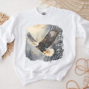 Puede incluir: Sudadera blanca con una ilustración detallada de un águila calva en vuelo. El águila tiene plumas marrones y blancas, con un fondo de bosque nevado. Cuello redondo.