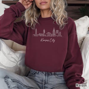 May include: Maroon-colored sweatshirt featuring a white line drawing of the Kansas City skyline. The words "Kansas City" are written in cursive below the skyline. The sweatshirt is a crew neck style.