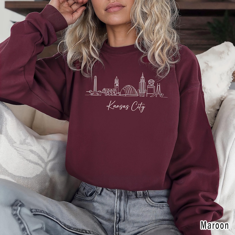 May include: Maroon-colored sweatshirt featuring a white line drawing of the Kansas City skyline. The words "Kansas City" are written in cursive below the skyline. The sweatshirt is a crew neck style.