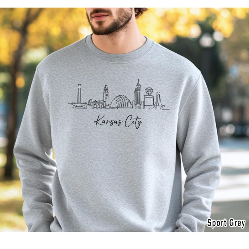 May include: A light grey sweatshirt featuring a black line drawing of the Kansas City skyline. The words "Kansas City" are written in a cursive font below the skyline. The bottom right corner of the sweatshirt reads "Sport Grey".