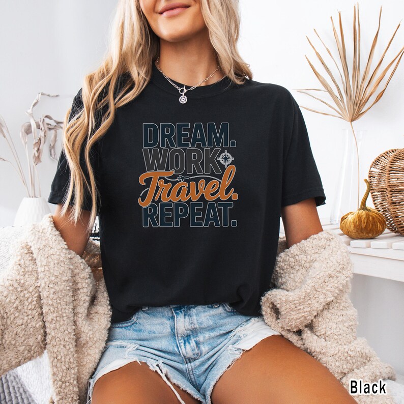 May include: Black t-shirt with the text "DREAM. WORK. Travel. REPEAT." in white and orange lettering. The shirt is a crew neck style and is being worn by a person.
