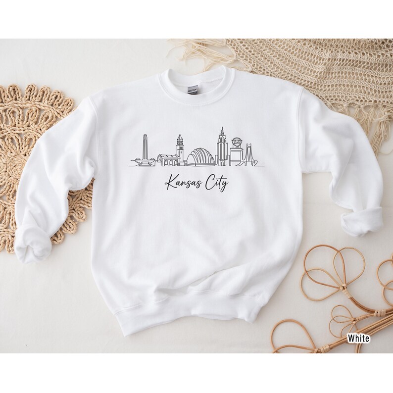 May include: White crewneck sweatshirt featuring a black line drawing of the Kansas City skyline. The words "Kansas City" are written in a script font below the skyline. The sweatshirt has a relaxed fit and long sleeves.