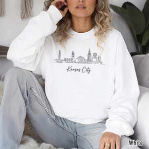 May include: A white ceramic mug with a black handle and the text "But First, Coffee" in a bold, black font. The mug is set against a blurred background, suggesting a cozy setting.