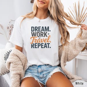 May include: White t-shirt with the text "DREAM. WORK. Travel. REPEAT." in black and orange lettering. The word "Travel" is in a cursive font. The shirt is a crew neck style.