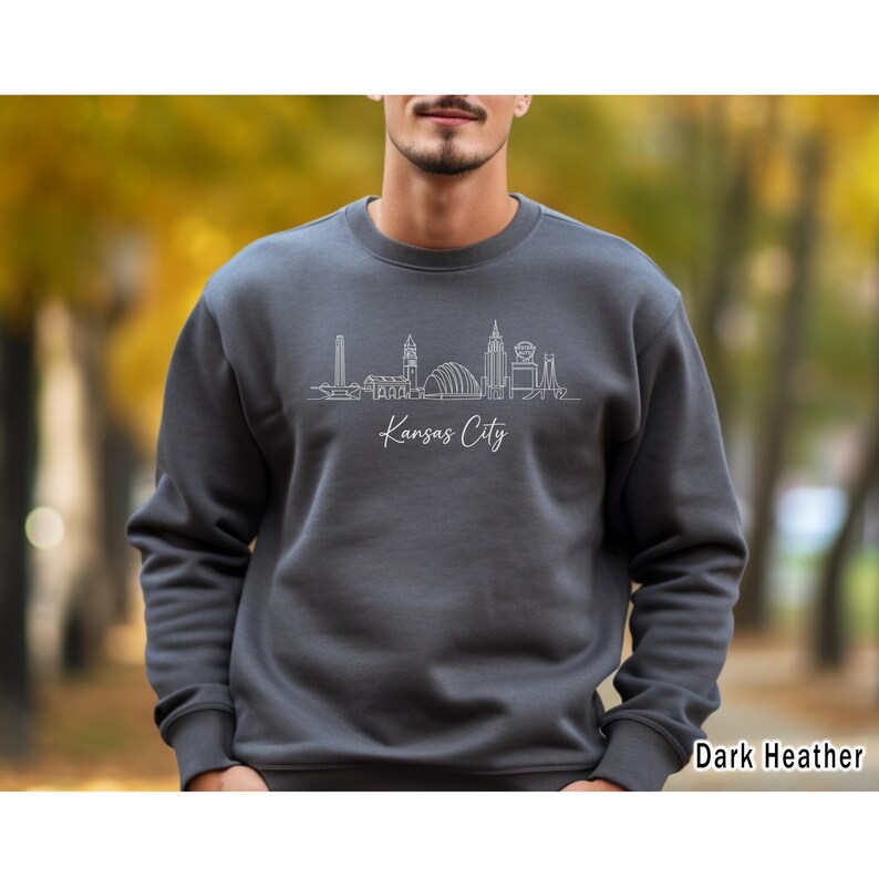 May include: Dark heather gray sweatshirt featuring a white line drawing of the Kansas City skyline. The city's name is written in a cursive font below the skyline. The sweatshirt is a crew neck style.