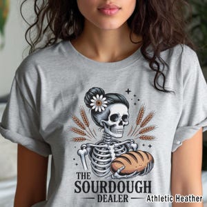 May include: Grey athletic heather t-shirt with a skeleton design holding bread, and a daisy in its hair. The text reads "THE SOURDOUGH DEALER". Wheat stalks are positioned around the skeleton. The t-shirt is a casual wear item.