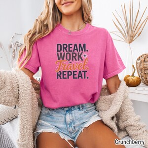 May include: A short-sleeved, pink t-shirt with the words "DREAM. WORK. Travel. REPEAT." in a layered design. The word "Travel" is in an orange script font, and the shirt is made of a soft material.