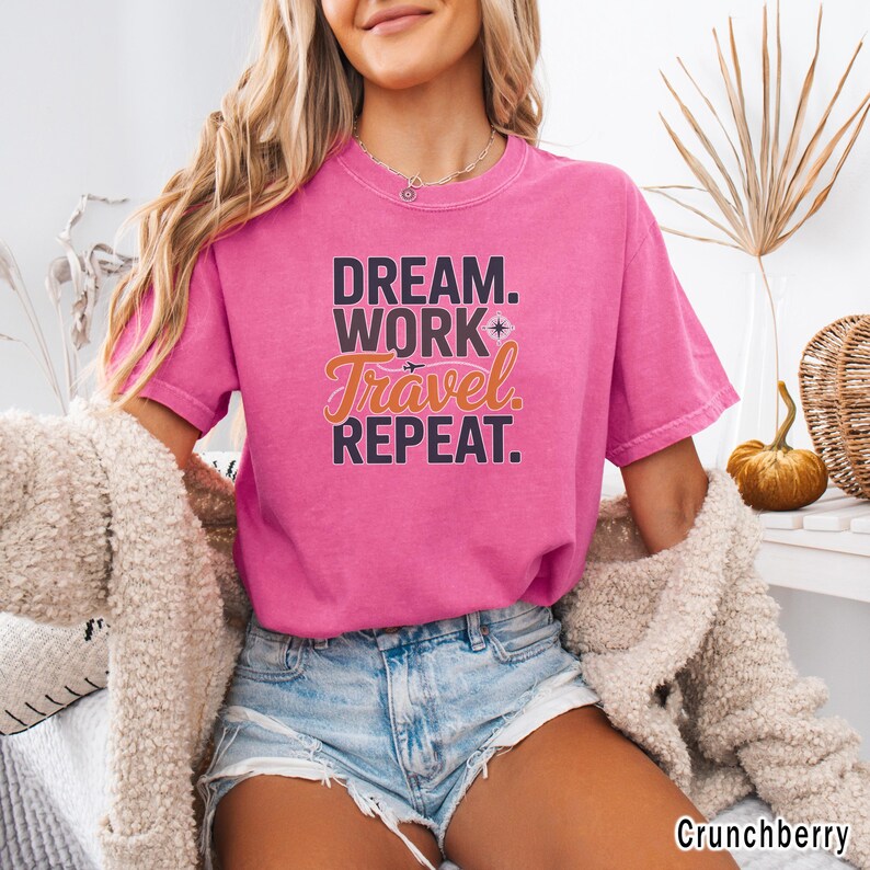 May include: A short-sleeved, pink t-shirt with the words "DREAM. WORK. Travel. REPEAT." in a layered design. The word "Travel" is in an orange script font, and the shirt is made of a soft material.