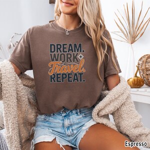 May include: Espresso-colored t-shirt with the text "DREAM. WORK. TRAVEL. REPEAT." in a mix of black and orange lettering. The shirt is a crew neck and has short sleeves. The model is wearing denim shorts and a cream-colored cardigan.