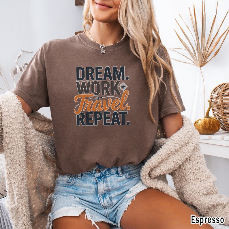 May include: Espresso-colored t-shirt with the text "DREAM. WORK. TRAVEL. REPEAT." in a mix of black and orange lettering. The shirt is a crew neck and has short sleeves. The model is wearing denim shorts and a cream-colored cardigan.