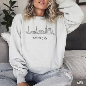 May include: Light gray sweatshirt with a black line drawing of the Kansas City skyline. The words "Kansas City" are written in a script font below the skyline. The sweatshirt is a crew neck style.