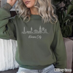 May include: Military green sweatshirt featuring a white line drawing of the Kansas City skyline. The words "Kansas City" are written in cursive below the skyline. The sweatshirt has ribbed cuffs and a ribbed hem.