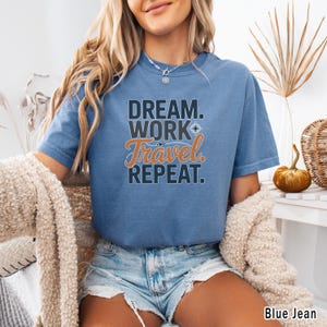 May include: Blue jean colored t-shirt with the text "DREAM. WORK. Travel. REPEAT." in black and orange. The shirt has short sleeves and a crew neck. The shirt is a comfortable, casual style.