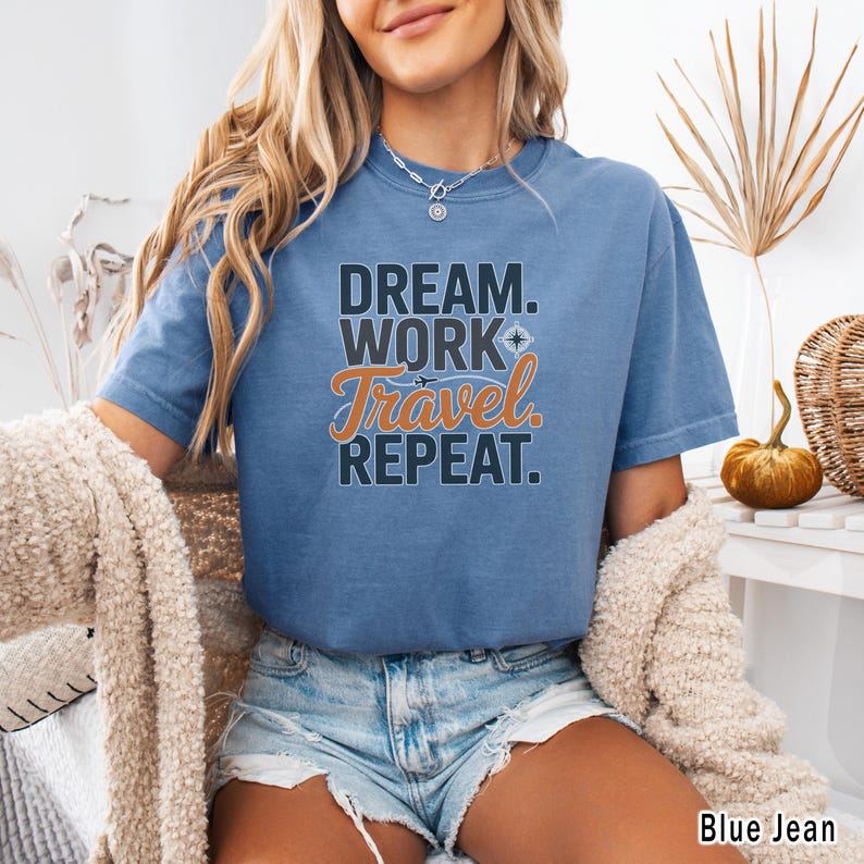 May include: Blue jean colored t-shirt with the text "DREAM. WORK. Travel. REPEAT." in black and orange. The shirt has short sleeves and a crew neck. The shirt is a comfortable, casual style.