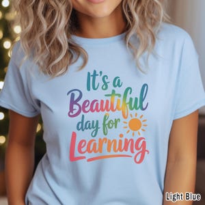 May include: Light blue t-shirt with the text "It's a Beautiful day for Learning" in a rainbow gradient. A small orange sun graphic is also included. The shirt is a light blue color.