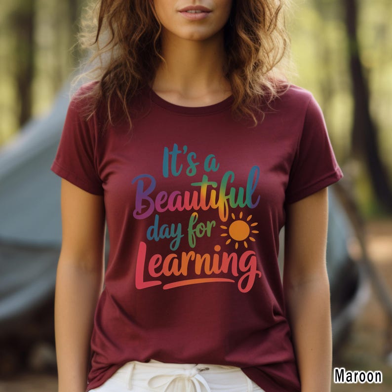 May include: Maroon t-shirt with the text "It's a Beautiful day for Learning" in a rainbow gradient. The design includes a sun graphic. The shirt is a short-sleeved crew neck.