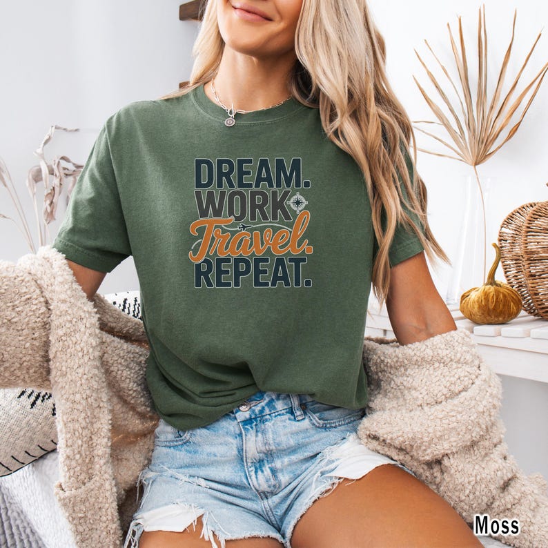 May include: A moss green t-shirt with the text "DREAM. WORK. TRAVEL. REPEAT." in a layered design. The word "Travel" is in orange script. The shirt is short-sleeved and has a relaxed fit.