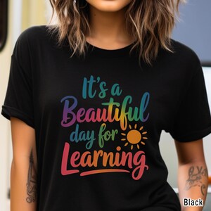 May include: Black t-shirt with the text "It's a Beautiful day for Learning" in a rainbow gradient. A yellow and orange sun graphic is included. The shirt is a crew neck style.