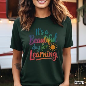 May include: Dark green t-shirt with the text "It's a Beautiful day for Learning" in rainbow colors, with a yellow sun graphic. The shirt is a short-sleeved crew neck.