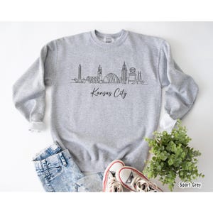 May include: A light gray sweatshirt with a black line drawing of the Kansas City skyline and the words "Kansas City" in a script font. The sweatshirt is paired with blue jeans and white sneakers.