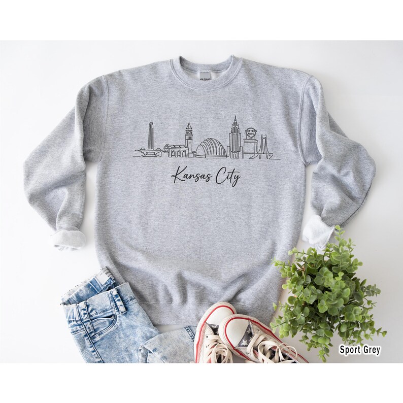 May include: A light gray sweatshirt with a black line drawing of the Kansas City skyline and the words "Kansas City" in a script font. The sweatshirt is paired with blue jeans and white sneakers.