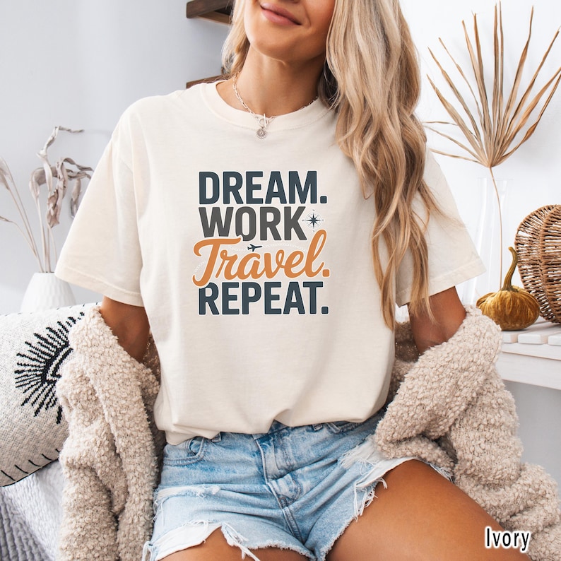 May include: Ivory-colored t-shirt with the text "DREAM. WORK. Travel. REPEAT." in black and orange. The shirt has short sleeves and a relaxed fit. The image shows a person wearing the shirt.