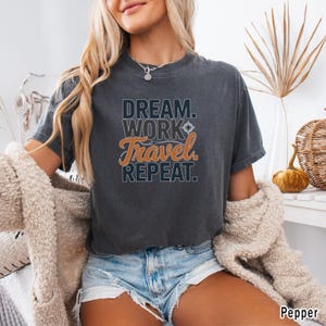 May include: Dark gray t-shirt with the words "DREAM. WORK. TRAVEL. REPEAT." in a mix of fonts and colors. The word "Travel" is in orange script. The shirt is a comfortable, casual style.