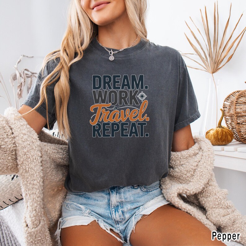 May include: Dark gray t-shirt with the words "DREAM. WORK. TRAVEL. REPEAT." in a mix of fonts and colors. The word "Travel" is in orange script. The shirt is a comfortable, casual style.