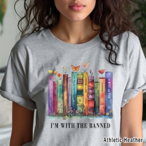 May include: A grey t-shirt featuring a watercolour illustration of a stack of banned books, including "Of Mice and Men", "Fahrenheit 451", "The Catcher in the Rye", and "The Handmaid's Tale". The text "I'm with the Banned" is printed below the illustration. The shirt is labelled "Athletic Heather".