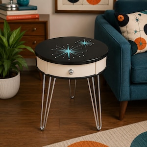 May include: A round side table with a black top featuring two light blue starburst designs. The table has a cream-colored drawer with a single silver knob and is supported by four thin, silver-toned metal hairpin legs.