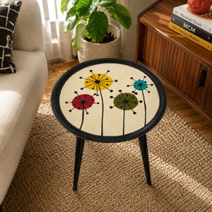 May include: A round side table with a black frame and three tapered black legs. The tabletop features a cream-colored surface decorated with a graphic pattern of four stylized dandelion-like flowers in yellow, red, olive green, and teal blue.