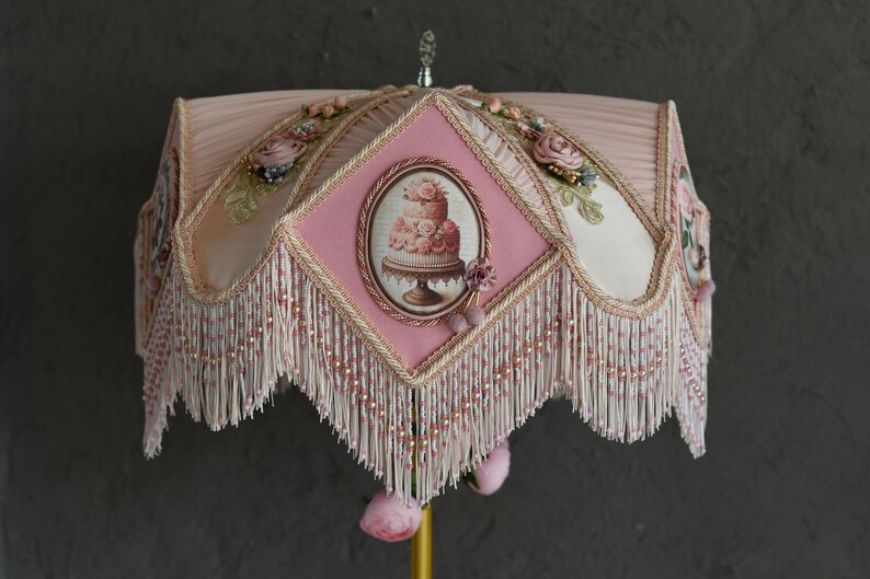 Pu&ograve; includere: Un paralume rosa con una frangia bianca e un motivo floreale con una torta. Il paralume ha un pannello centrale a forma di diamante con un bordo dorato.