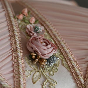 Pu&ograve; includere: Primo piano di un tessuto rosa con bordino dorato e bianco. Il tessuto &egrave; decorato con una rosa rosa e ricami dorati.