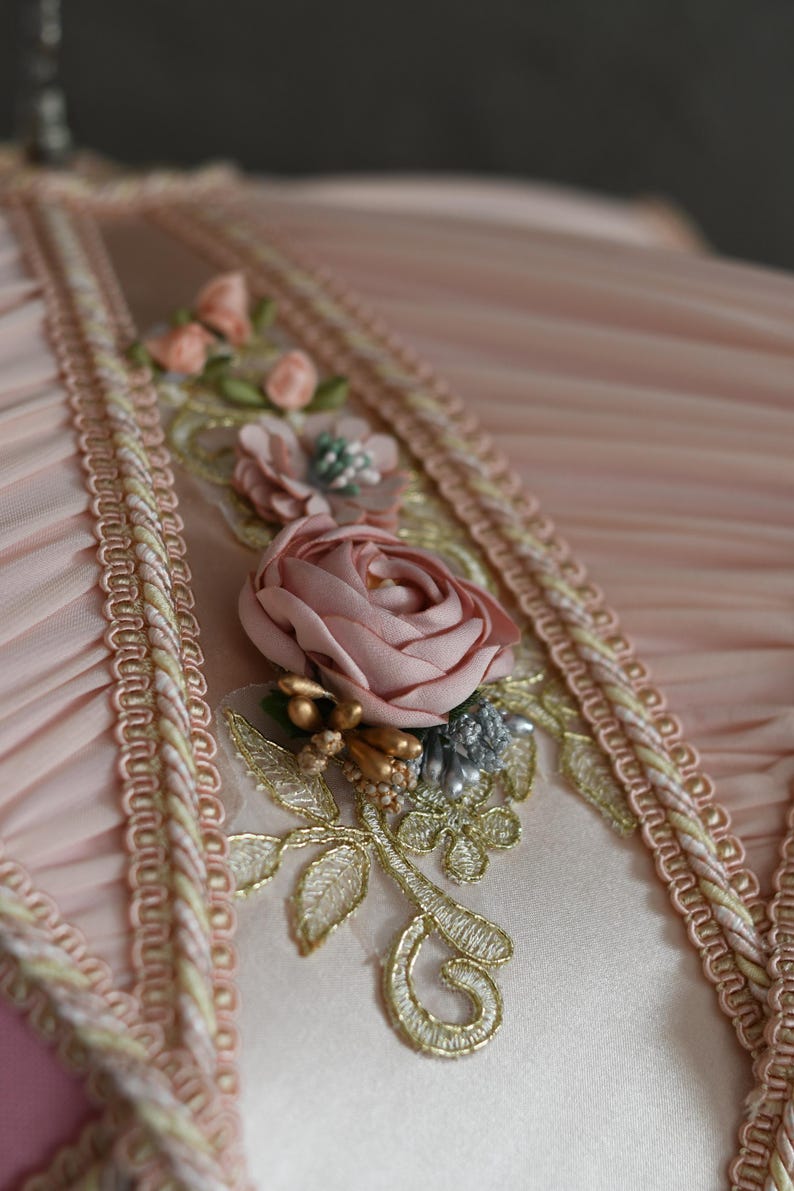 Pu&ograve; includere: Primo piano di un tessuto rosa con bordino dorato e bianco. Il tessuto &egrave; decorato con una rosa rosa e ricami dorati.